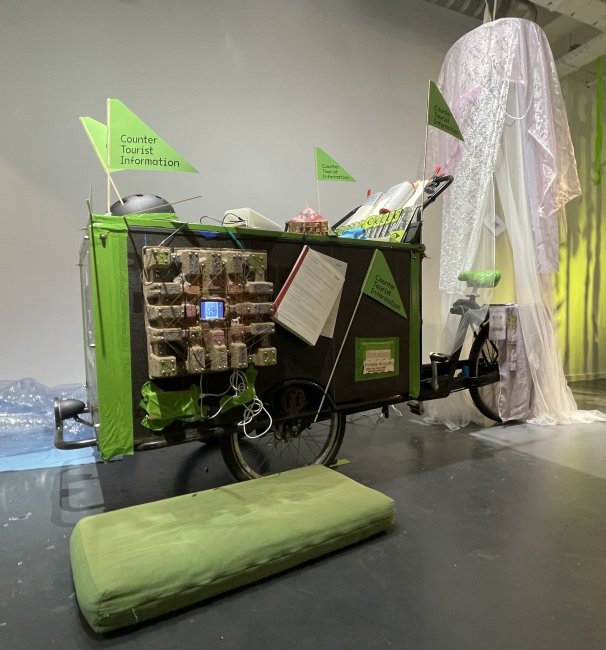 A funky looking cargo bike with small triangular flags attached saying "Counter Tourist Information" and with a whole collection of things attached, small computer screens, mosquito net, small booklets... Although It is parked in a gallery space, it is designed to make a temporary stand in the street.