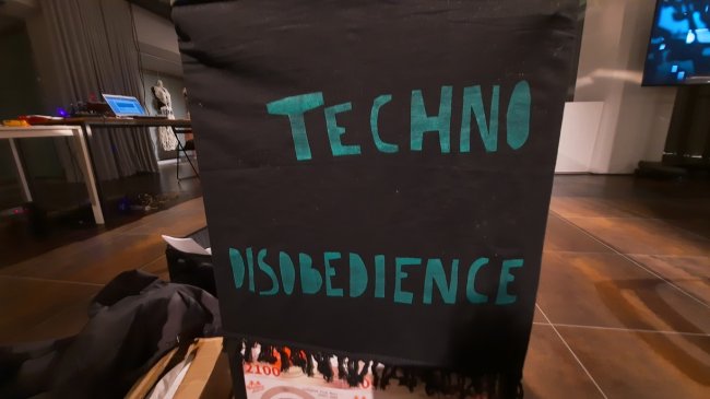 A black banner with the word Techno-disobedience wrote in green capital letter is held in front of the camera. In the background an exhibition space.