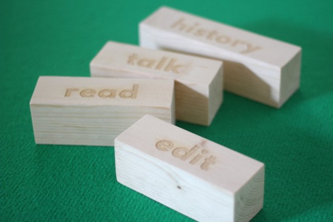 Four wooden blocks on a green carpet. Each of them carries an engraved word : "history", "talk", "read", "edit".