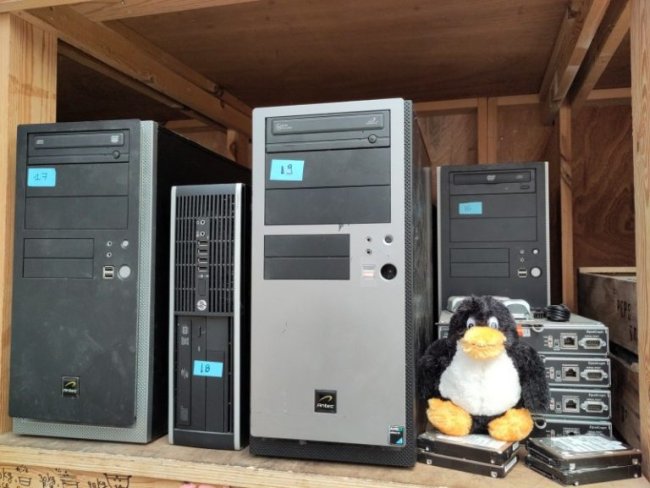 Several old computers and a teddy bear in the shape of a pinguin on a wodden shelf.