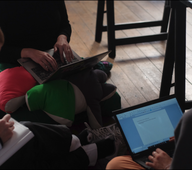 Part of 3 people sitting in a circle on the floor. Two people are working on portable computers while a third person is writing on a note book.